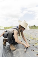 A person wearing a wide-brimmed hat and sunglasses is kneeling on a farm field, planting seedlings. They are dressed in work attire, including boots and overalls. The field has rows of small plants with holes prepared for planting, and the sky is partly cloudy.