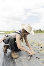 A person wearing a wide-brimmed hat and sunglasses is kneeling on a farm field, planting seedlings. They are dressed in work attire, including boots and overalls. The field has rows of small plants with holes prepared for planting, and the sky is partly cloudy.