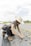 A person wearing a wide-brimmed hat and sunglasses is kneeling on a farm field, planting seedlings. They are dressed in work attire, including boots and overalls. The field has rows of small plants with holes prepared for planting, and the sky is partly cloudy.