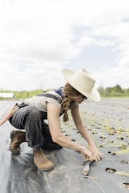 A person wearing a wide-brimmed hat and sunglasses is kneeling on a farm field, planting seedlings. They are dressed in work attire, including boots and overalls. The field has rows of small plants with holes prepared for planting, and the sky is partly cloudy.