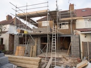 A house is under construction with scaffolding erected around the structure. The building appears to be a residential property being extended or renovated. Materials such as bricks and wooden planks are scattered in the foreground. Several ladders are visible, and the windows of the house have been removed or are in the process of being installed.
