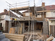 A house is under construction with scaffolding erected around the structure. The building appears to be a residential property being extended or renovated. Materials such as bricks and wooden planks are scattered in the foreground. Several ladders are visible, and the windows of the house have been removed or are in the process of being installed.