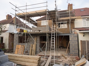 A house is under construction with scaffolding erected around the structure. The building appears to be a residential property being extended or renovated. Materials such as bricks and wooden planks are scattered in the foreground. Several ladders are visible, and the windows of the house have been removed or are in the process of being installed.