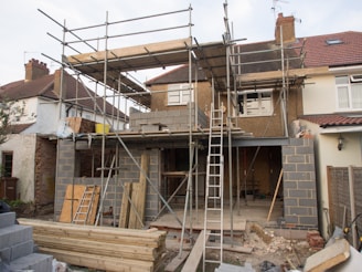 A house is under construction with scaffolding erected around the structure. The building appears to be a residential property being extended or renovated. Materials such as bricks and wooden planks are scattered in the foreground. Several ladders are visible, and the windows of the house have been removed or are in the process of being installed.