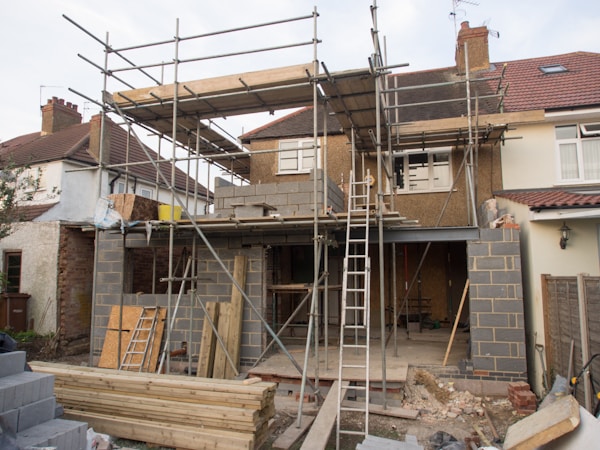 A house is under construction with scaffolding erected around the structure. The building appears to be a residential property being extended or renovated. Materials such as bricks and wooden planks are scattered in the foreground. Several ladders are visible, and the windows of the house have been removed or are in the process of being installed.