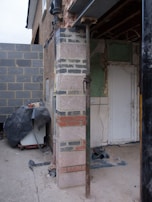 A partially constructed or renovated building area featuring exposed brickwork and construction materials. The image shows a corner pillar with different types of bricks and concrete, surrounded by a mixture of unfinished surfaces. There is a temporary metal support beam next to the pillar, and construction equipment is covered with a tarp. Exposed wiring and insulation can be seen on the inner wall, along with a door leading to another space.