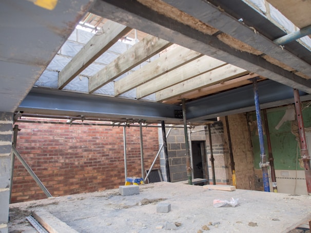 An unfinished interior construction site featuring exposed wooden beams and steel joists. The walls are comprised of brickwork, with some areas having bare insulation. Temporary support beams are holding up the structure, and construction materials are strewn across the dusty floor.