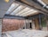 An unfinished interior construction site featuring exposed wooden beams and steel joists. The walls are comprised of brickwork, with some areas having bare insulation. Temporary support beams are holding up the structure, and construction materials are strewn across the dusty floor.