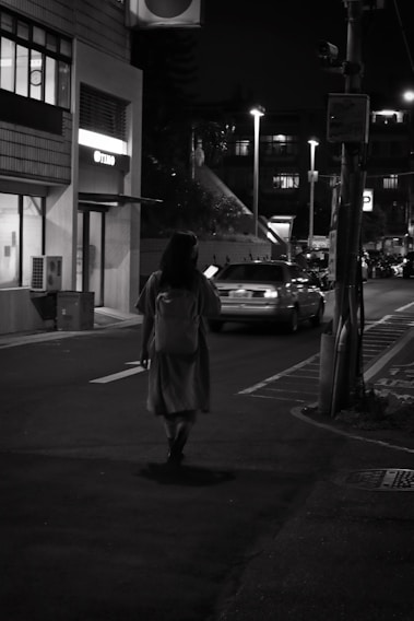 A woman walks alone on a dimly lit urban street at night. She is carrying a shoulder bag and is illuminated by the soft glow of streetlights. Nearby, cars are parked along the roadside, and a building with lit windows is visible. The atmosphere is quiet and contemplative.
