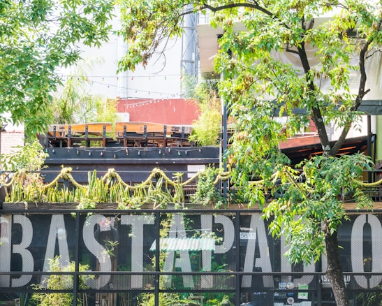 A lush garden setting features prominently with greenery, including trees and hanging plants, partially obscuring a modern building. The building's facade has a decorative mesh with large lettering. The area is well-lit by natural light, and there's an inviting atmosphere with outdoor seating visible above.