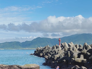 A coastal landscape featuring large geometric concrete wave breakers extending into the sea, with a bright red lighthouse positioned prominently among them. In the background, a range of forested mountains under a partly cloudy blue sky creates a picturesque scene. A person wearing a pink hat is visible near the foreground on the right side.
