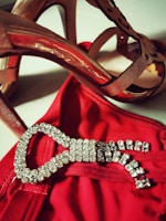 High-heeled shoes in a brownish shade are positioned above a red garment adorned with a sparkling, jewel-encrusted buckle.