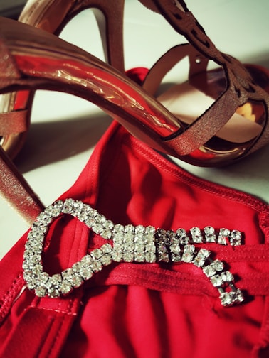 High-heeled shoes in a brownish shade are positioned above a red garment adorned with a sparkling, jewel-encrusted buckle.