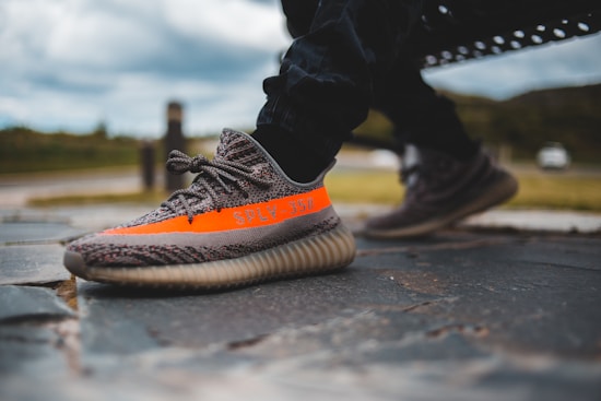 A close-up of a pair of stylish sneakers with a prominent orange stripe and text, worn by a person sitting outdoors. The setting appears to be a park or an open area with a stone-paved ground.
