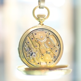 An intricate gold pocket watch with visible gears and mechanisms, showcasing detailed craftsmanship. The watch is open to reveal its inner workings with small gears, screws, and jewels that are meticulously arranged. The background is softly blurred, emphasizing the watch's complexity and elegance.