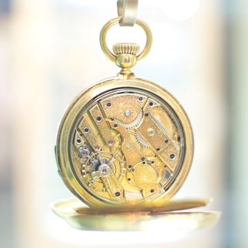 An intricate gold pocket watch with visible gears and mechanisms, showcasing detailed craftsmanship. The watch is open to reveal its inner workings with small gears, screws, and jewels that are meticulously arranged. The background is softly blurred, emphasizing the watch's complexity and elegance.