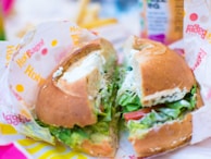 A close-up of a bagel sandwich filled with various fresh ingredients such as lettuce, tomato, and cream cheese. The sandwich is set on a colorful wrapper with 'Holy Bagel' branding visible. The background shows blurred elements, possibly a drink and fries.