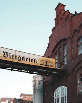 A large brick building with intricate architectural details is pictured. Prominently displayed is a sign with the word 'Biergarten,' suggesting a beer garden area. The architectural style of the building appears to be historical, with arched windows and a rusted metal cylinder in the foreground.