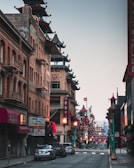 A quiet street scene in Chinatown featuring traditional architecture with pagoda-style roofs. The buildings are adorned with red and gold accents, and there are street lamps with decorative designs. Lanterns are hung across the street, creating a festive atmosphere. Several cars are parked along the street, and the area seems calm with no visible pedestrians.
