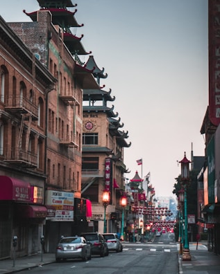 A quiet street scene in Chinatown featuring traditional architecture with pagoda-style roofs. The buildings are adorned with red and gold accents, and there are street lamps with decorative designs. Lanterns are hung across the street, creating a festive atmosphere. Several cars are parked along the street, and the area seems calm with no visible pedestrians.