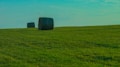 A serene landscape featuring two large, cylindrical hay bales positioned on a vast green field under a clear blue sky. The grass appears lush and well-kept, stretching out to the horizon.