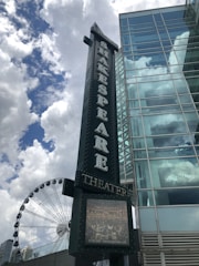 A vertical signboard with the word 'SHAKESPEARE THEATER' in illuminated letters is prominent in the foreground. Adjacent to it is a modern glass building with reflective windows. In the background, partially obscured, there is a Ferris wheel and a sky filled with white fluffy clouds against a bright blue backdrop.