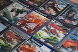 A collection of PlayStation 2 video game cases arranged closely together, featuring titles such as Ace Combat and Burnout. The cases display a mix of action and racing game covers, with vibrant images of fighter jets and cars.