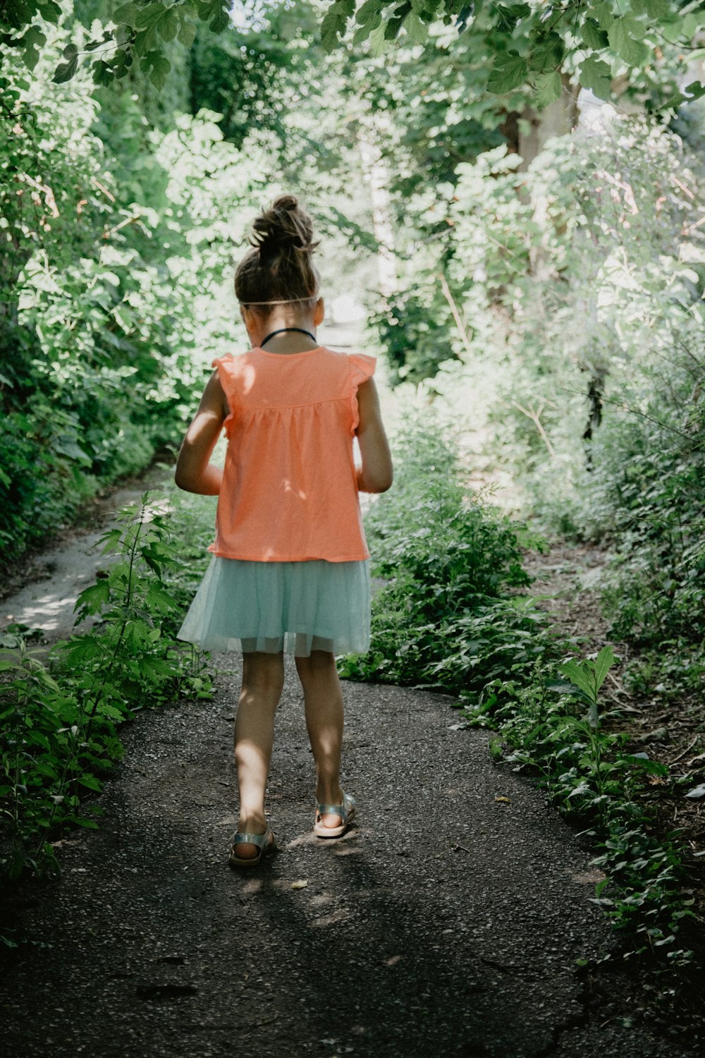 ピンクのタンクトップとブルーデニムのショートパンツを着た女性が、緑の植物の間の小道に立っています。