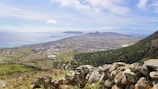 A scenic landscape featuring a coastal town with sprawling fields and forests, leading to the sea. The horizon meets a mountain range in the distance, and fluffy clouds are scattered across a blue sky. A foreground of rugged stone adds texture to the composition.