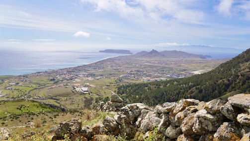 A scenic landscape featuring a coastal town with sprawling fields and forests, leading to the sea. The horizon meets a mountain range in the distance, and fluffy clouds are scattered across a blue sky. A foreground of rugged stone adds texture to the composition.
