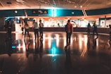 A group of people roller skating indoors, under warm and colorful lighting. The setting resembles a retro-themed roller rink with reflective flooring and a concession area in the background. The ceiling has an arrangement of lights that cast reflections on the floor, creating an inviting and lively atmosphere.