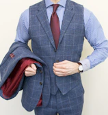 A person is wearing a stylish three-piece suit consisting of a blue checkered vest and pants with a matching jacket draped over the arm. The shirt underneath is blue and white striped, and the ensemble is completed with a dark red tie. A classic watch is visible on the wrist.