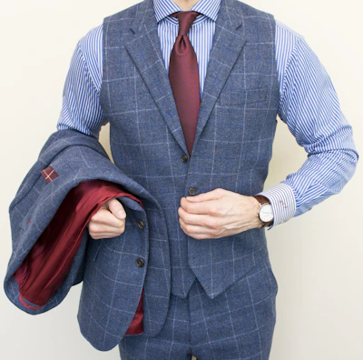 A person is wearing a stylish three-piece suit consisting of a blue checkered vest and pants with a matching jacket draped over the arm. The shirt underneath is blue and white striped, and the ensemble is completed with a dark red tie. A classic watch is visible on the wrist.