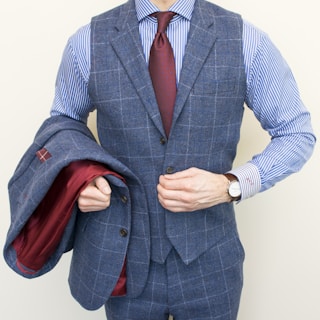 A person is wearing a stylish three-piece suit consisting of a blue checkered vest and pants with a matching jacket draped over the arm. The shirt underneath is blue and white striped, and the ensemble is completed with a dark red tie. A classic watch is visible on the wrist.