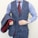 A person is wearing a stylish three-piece suit consisting of a blue checkered vest and pants with a matching jacket draped over the arm. The shirt underneath is blue and white striped, and the ensemble is completed with a dark red tie. A classic watch is visible on the wrist.