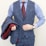 A person is wearing a stylish three-piece suit consisting of a blue checkered vest and pants with a matching jacket draped over the arm. The shirt underneath is blue and white striped, and the ensemble is completed with a dark red tie. A classic watch is visible on the wrist.