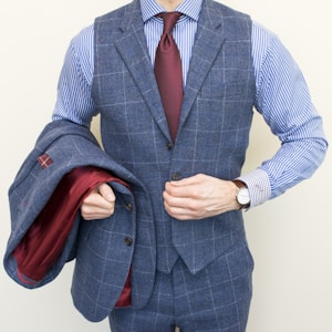 A person is wearing a stylish three-piece suit consisting of a blue checkered vest and pants with a matching jacket draped over the arm. The shirt underneath is blue and white striped, and the ensemble is completed with a dark red tie. A classic watch is visible on the wrist.
