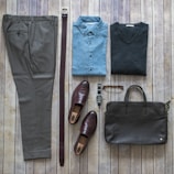 A neatly arranged flat lay of men's clothing and accessories on a wooden surface. The items include a pair of brown checked trousers, a dark brown belt, a blue button-up shirt, a dark grey sweater, brown loafers, a wristwatch with a metal strap, a pair of brown eyeglasses, and a dark brown leather briefcase.