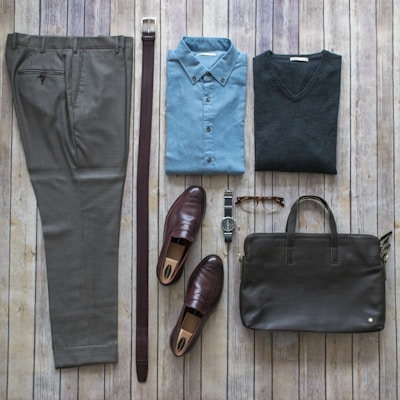 A neatly arranged flat lay of men's clothing and accessories on a wooden surface. The items include a pair of brown checked trousers, a dark brown belt, a blue button-up shirt, a dark grey sweater, brown loafers, a wristwatch with a metal strap, a pair of brown eyeglasses, and a dark brown leather briefcase.