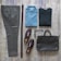 A neatly arranged flat lay of men's clothing and accessories on a wooden surface. The items include a pair of brown checked trousers, a dark brown belt, a blue button-up shirt, a dark grey sweater, brown loafers, a wristwatch with a metal strap, a pair of brown eyeglasses, and a dark brown leather briefcase.