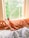 A cozy bed is positioned next to a window with orange silk pillows and a white comforter. A small potted plant rests on the windowsill. Sunlight casts warm tones onto the scene, and lush green trees are visible outside the window.