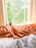 A cozy bed is positioned next to a window with orange silk pillows and a white comforter. A small potted plant rests on the windowsill. Sunlight casts warm tones onto the scene, and lush green trees are visible outside the window.