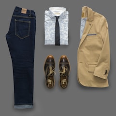 An outfit consisting of dark denim jeans, a light patterned shirt with a dark tie, a beige blazer, and brown leather shoes laid out flat. The blazer features a pocket square.