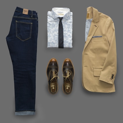 An outfit consisting of dark denim jeans, a light patterned shirt with a dark tie, a beige blazer, and brown leather shoes laid out flat. The blazer features a pocket square.