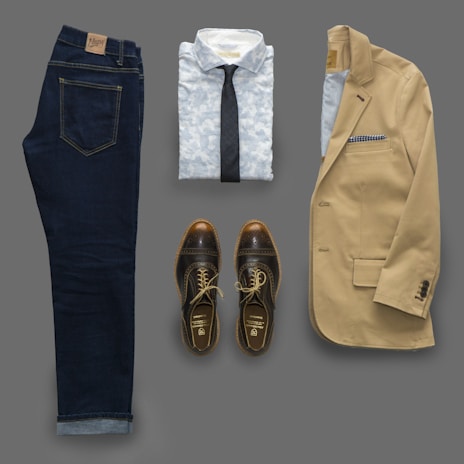 An outfit consisting of dark denim jeans, a light patterned shirt with a dark tie, a beige blazer, and brown leather shoes laid out flat. The blazer features a pocket square.