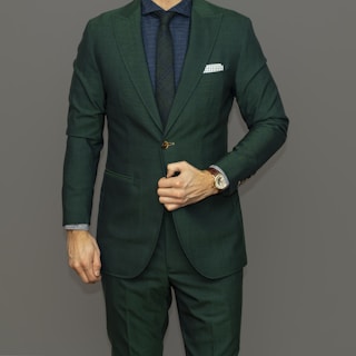 A well-dressed person is wearing a dark green suit paired with a navy blue shirt and matching tie. A white pocket square is tucked into the breast pocket. The person is adjusting the suit jacket with one hand, revealing a watch on the wrist.
