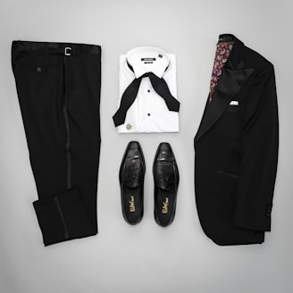 A neatly arranged flat lay of formal menswear, including a black suit jacket with satin lapels, matching black trousers, a white dress shirt with a black bow tie, and a pair of black leather dress shoes.