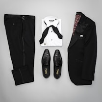 A neatly arranged flat lay of formal menswear, including a black suit jacket with satin lapels, matching black trousers, a white dress shirt with a black bow tie, and a pair of black leather dress shoes.