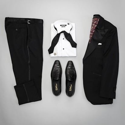 A neatly arranged flat lay of formal menswear, including a black suit jacket with satin lapels, matching black trousers, a white dress shirt with a black bow tie, and a pair of black leather dress shoes.