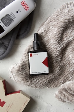 A bottle of CBD oil with a dropper lies on a textured, fluffy beige blanket. An electric device labeled SAFR is positioned to the top left, alongside the blanket. A partially visible cardboard box with red and white design is on the lower left corner.